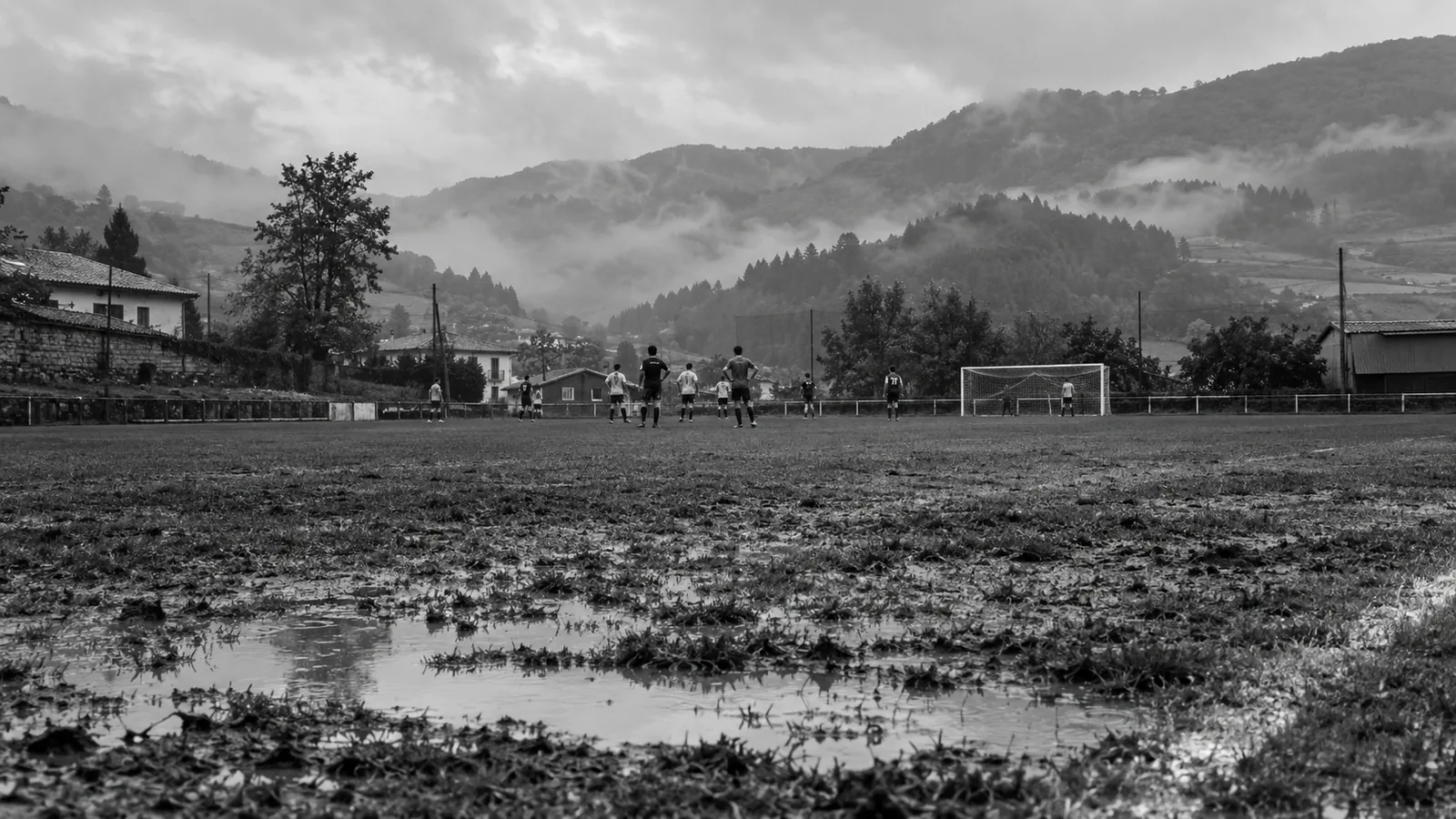Black-and-white image of a rainy neighborhood soccer field with players and misty hills.